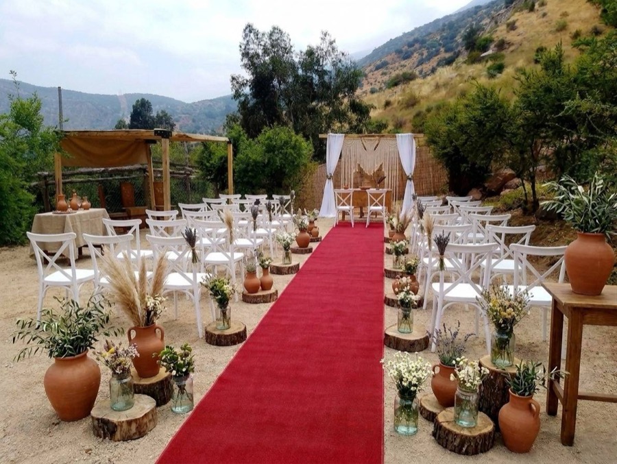 Pasillo de ceremonia con alfombra roja, sillas blancas y pampas, mirando al cerro